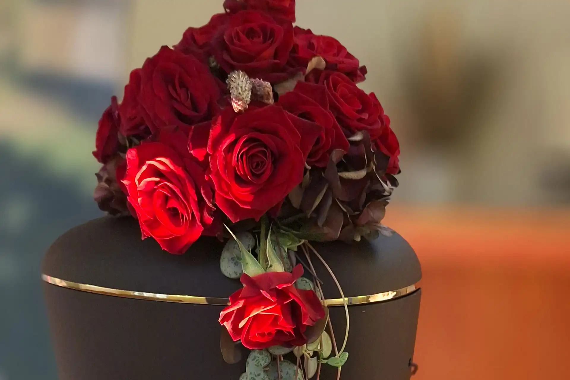 Greta - Blumen, Ideen & mehr... Inspiriert von der Liebe zur Natur. Blumen für besondere Momente & Floristik für jeden Anlass - Trauer Wir beraten Sie gern. Rote Rosen auf schwarzer Urne mit goldfarbenem Dekor.